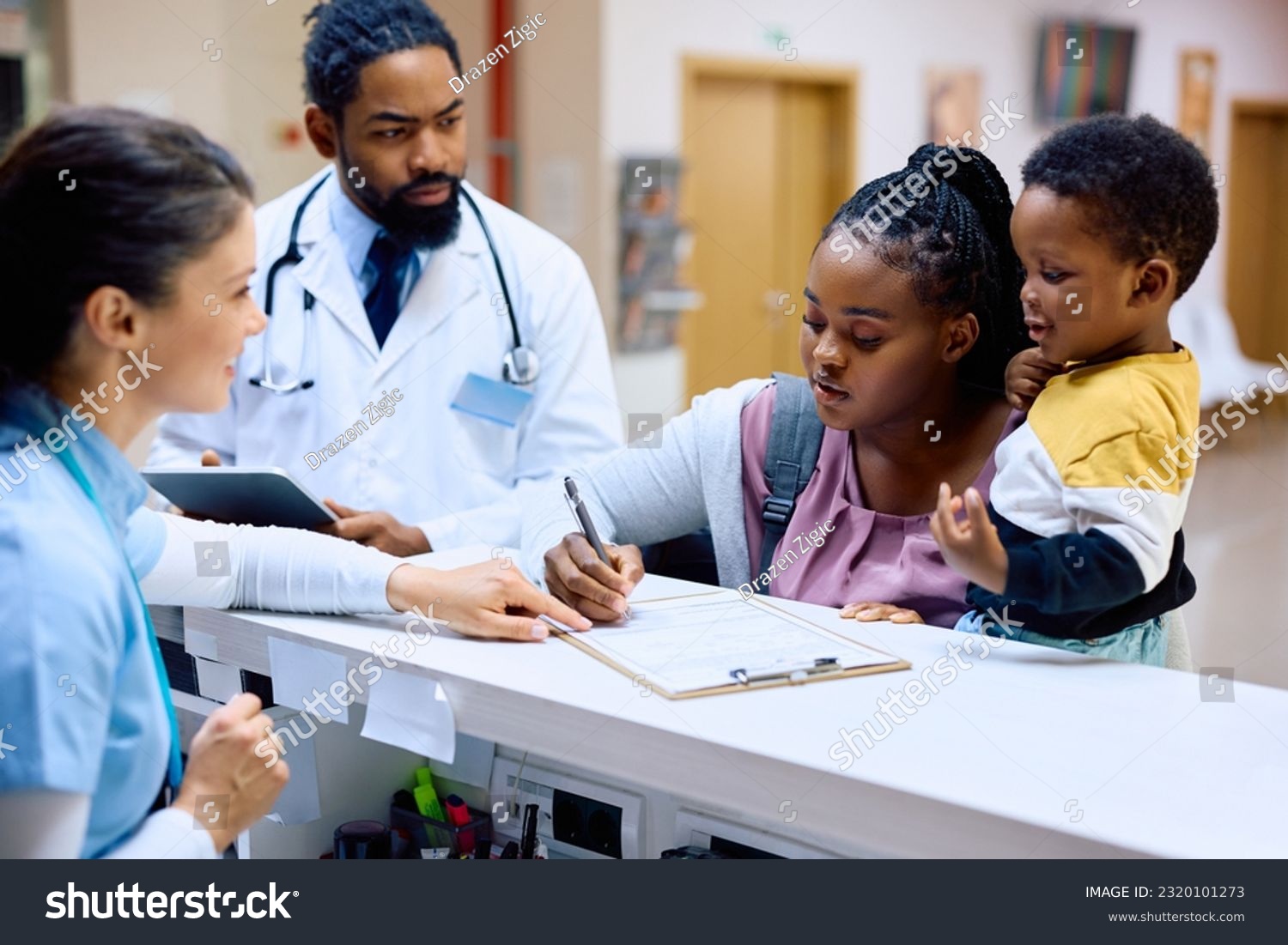 African American mother holding her small son while filling medical paperwork at reception desk in the hospital. #2320101273