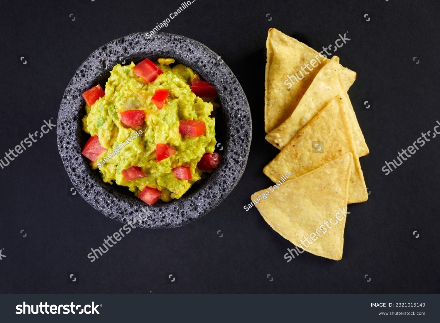 Top view of a delicious avocado guacamole with red diced tomatoes in a typical stone Mexican mortar or molcajete by a few totopos or nachos over a black background. #2321015149