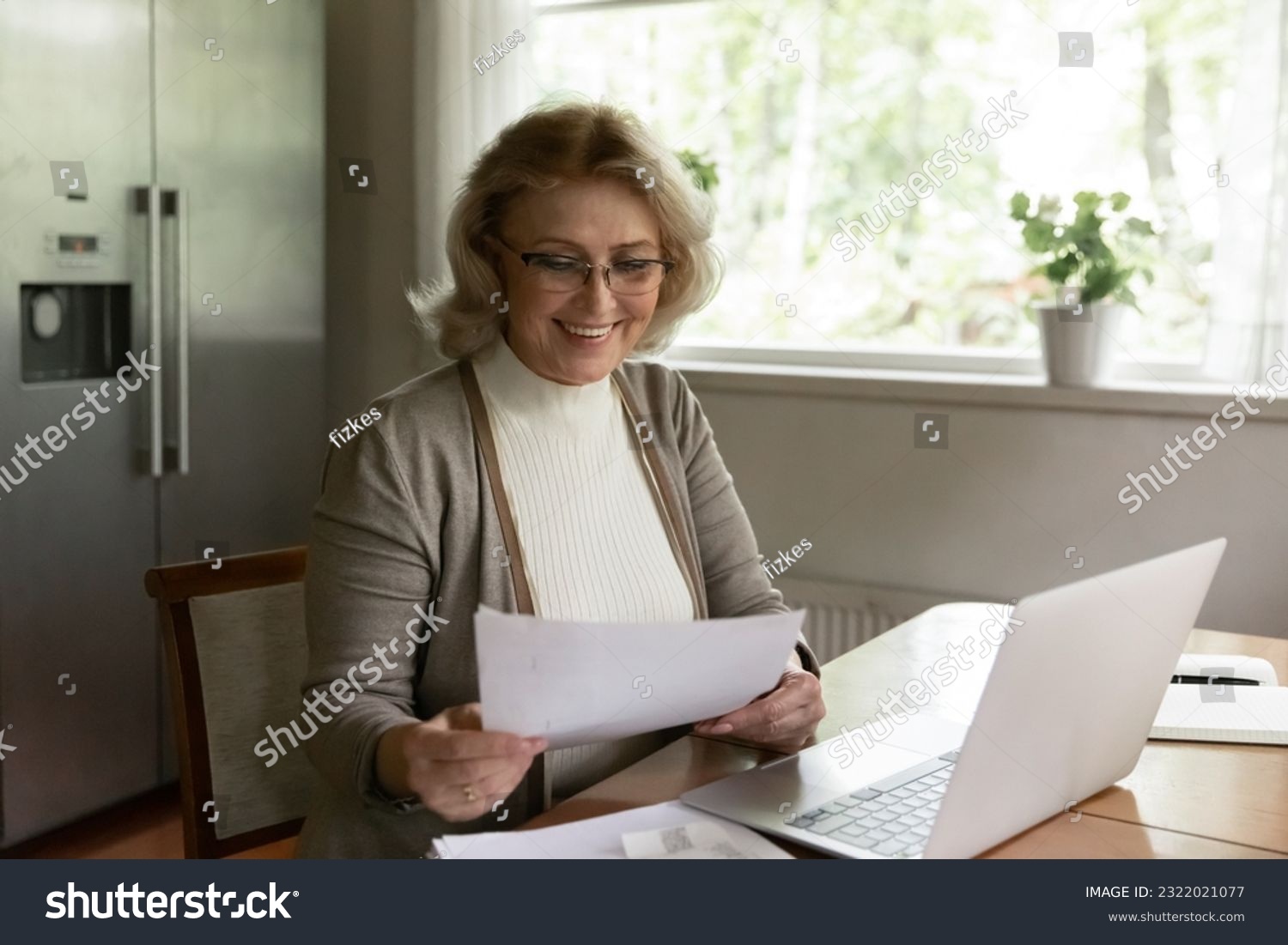 Smiling mature Caucasian 60s woman sit at desk at home work on computer with paperwork correspondence. Happy old female busy with paper documents pay bills online use modern laptop. #2322021077