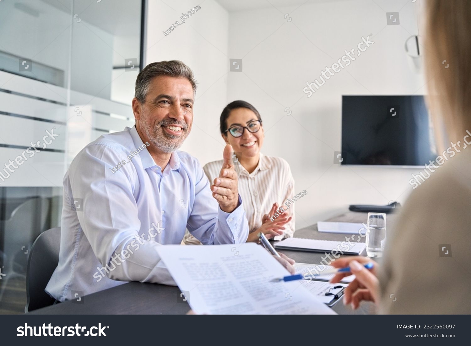Smiling Latin manager working with diverse colleagues at team meeting. Happy diverse business people international corporate executives talking at group briefing, collaborating at boardroom table. #2322560097