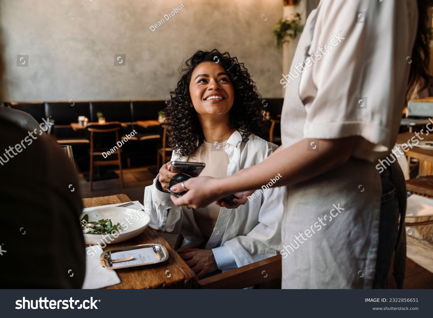 Young smiling african woman using smartphone while paying restaurant bill with contactless payment during dinner party with friends #2322856651