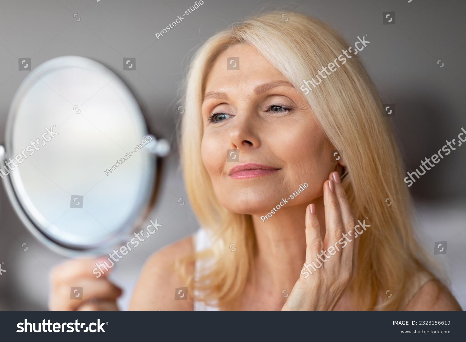 Attractive aged european woman looking in magnifying mirror and touching her face smooth skin, beautiful senior lady enjoying her appearance, closeup #2323156619