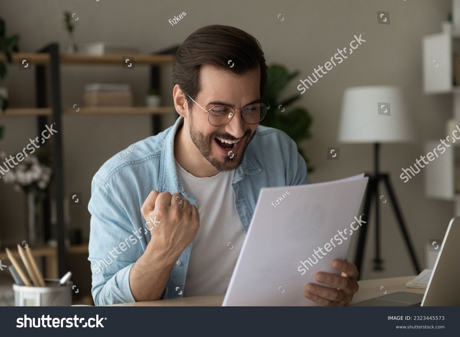 Overjoyed young man in eyeglasses looking through paper document, feeling excited of reading amazing news in letter celebrating getting lottery win notification or bank mortgage loan approval. #2323445573