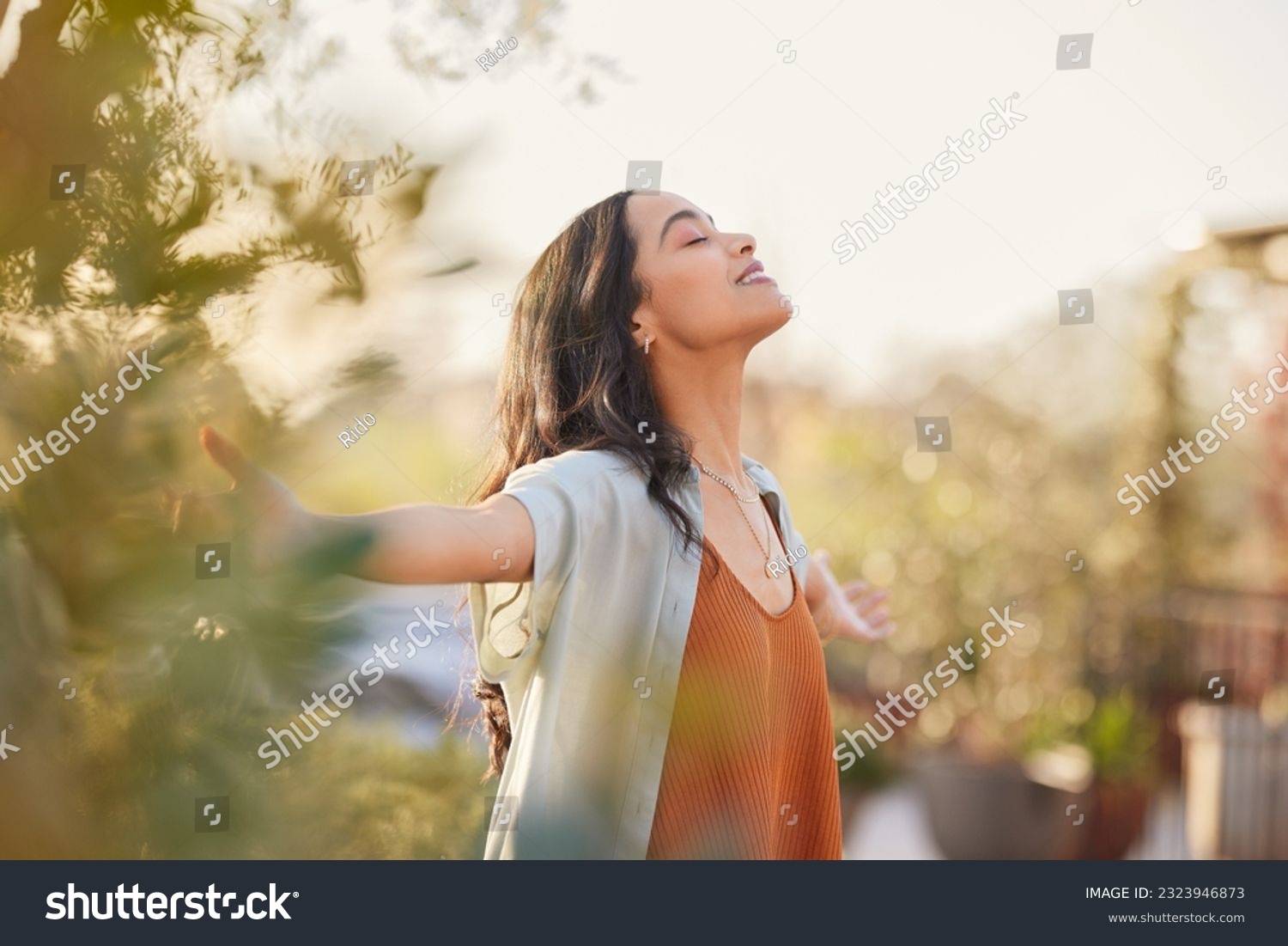Young latin woman with arms outstretched breathing in fresh air during sunrise at the balcony. Girl enjoying nature while meditating during morning with open arms and closed eyes. Mindful woman relax. #2323946873