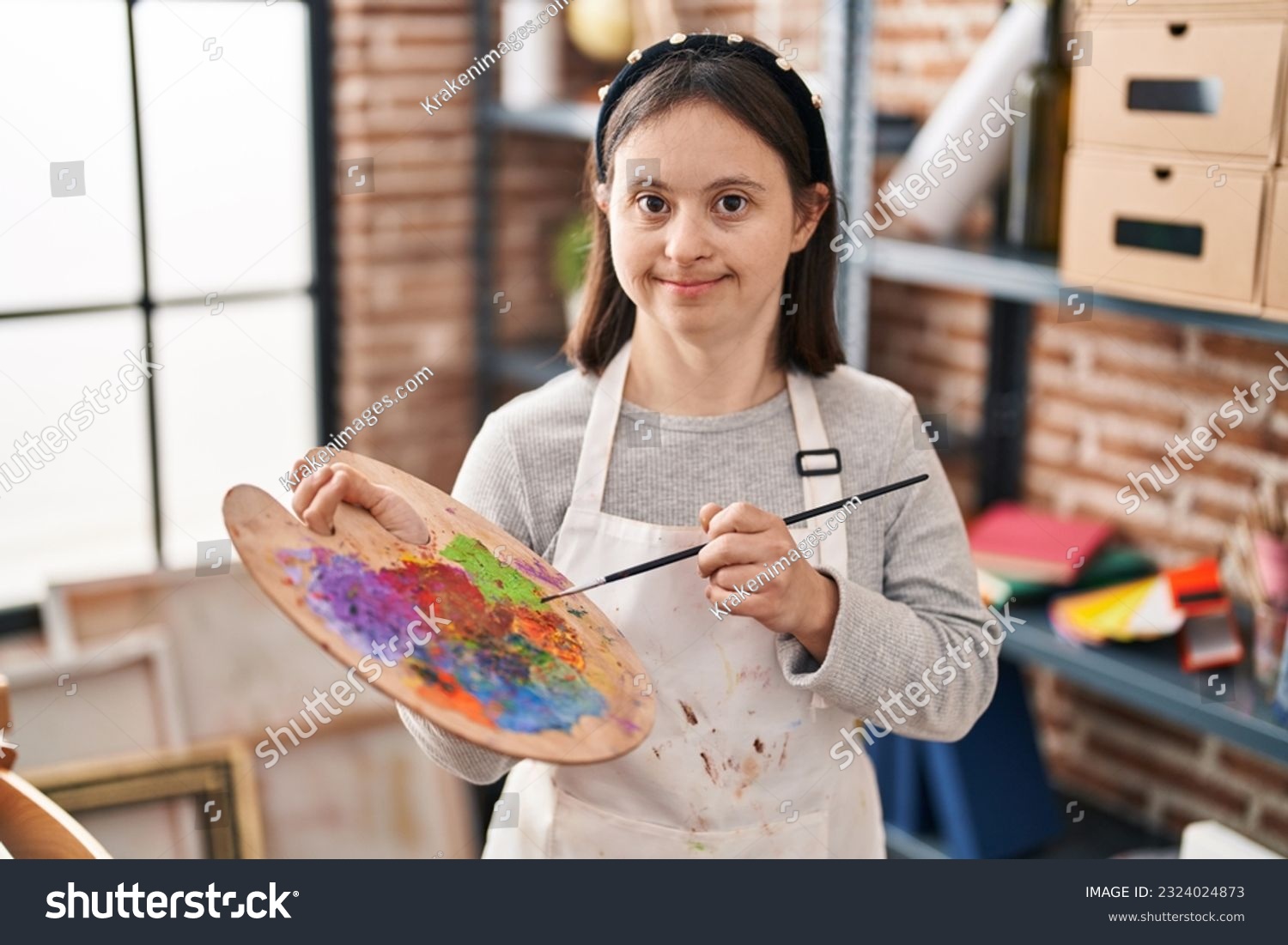 Young woman with down syndrome smiling confident holding paintbrush and palette at art studio #2324024873