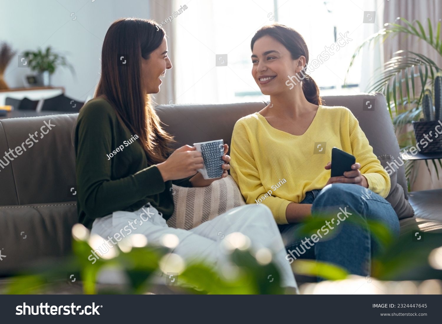 Shot of two smiling young women talking while drinking coffee sitting on couch in the living room at home. #2324447645