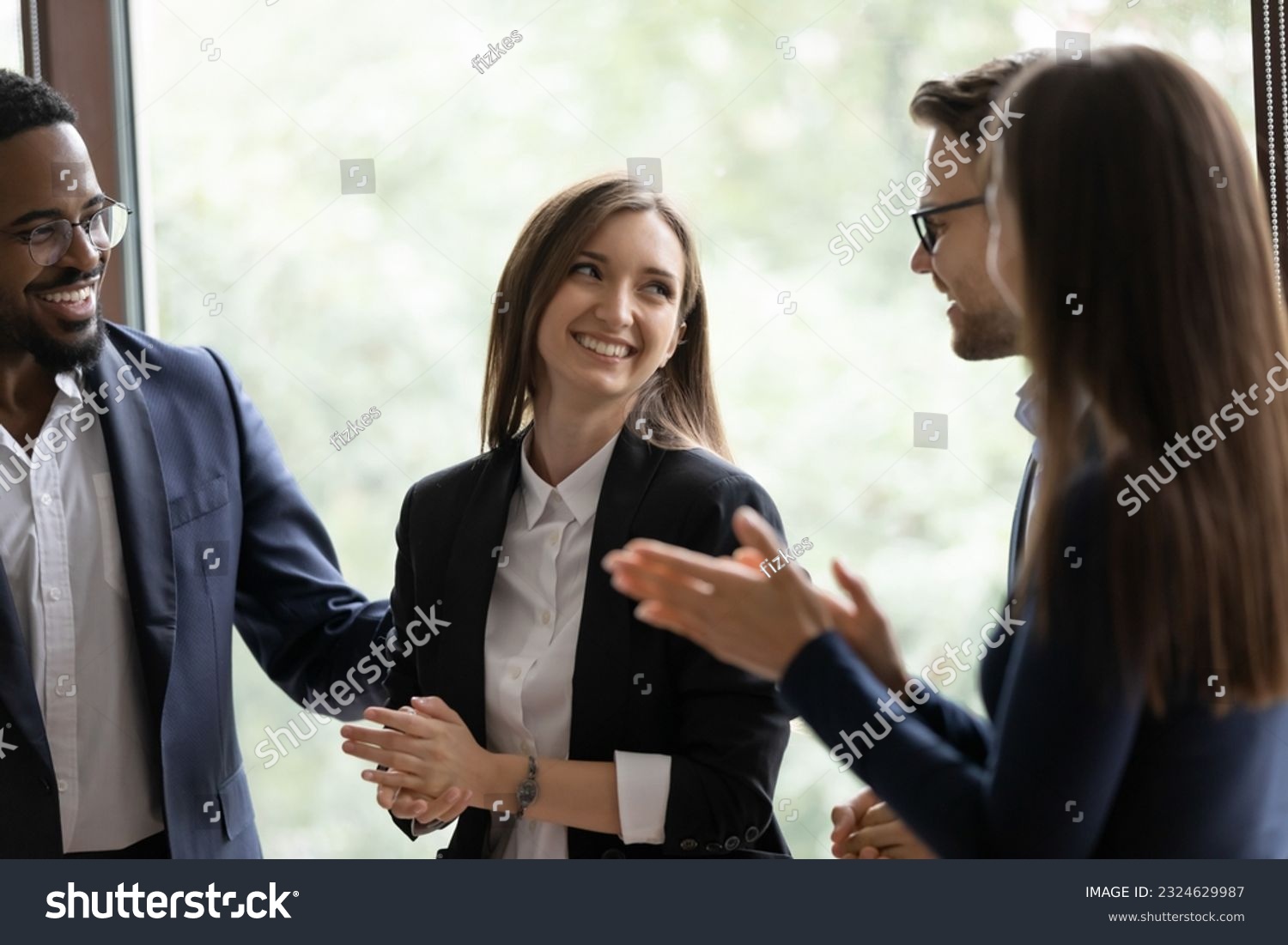 Laughing employees warmly welcoming young lady intern on workplace, friendly teammates congratulating woman coworker with promotion, partners praising businesswoman for creating successful project #2324629987