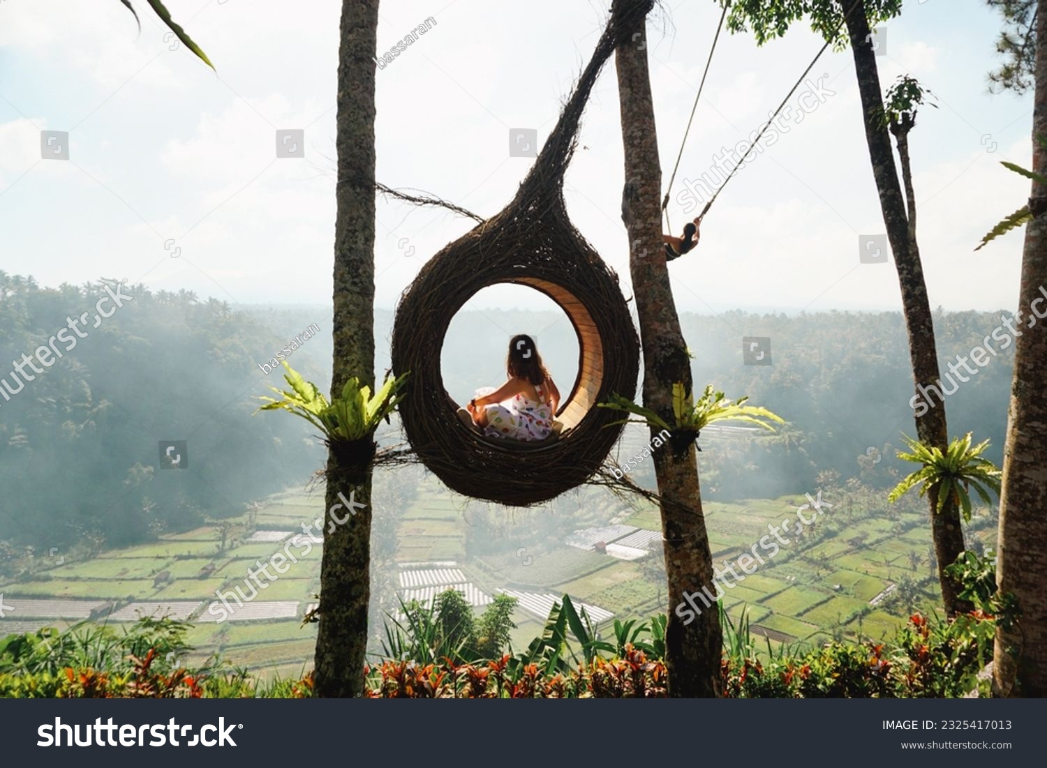 A young woman tourist sitting in a bird nest, immersed in the breathtaking green landscape of Bali on a sunny day. Tourist doing jungle swing. Rice fields and forest in background. Indonesia. #2325417013
