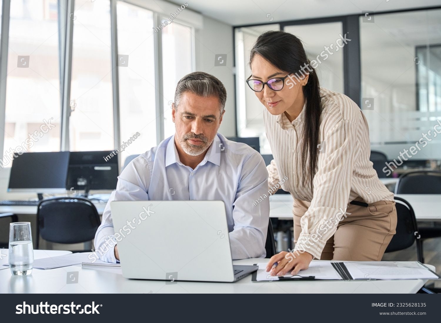 Two busy diverse professional coworkers discussing work using laptop in office. Asian employee learning online project discussing business plan with mature manager looking at computer at meeting. #2325628135