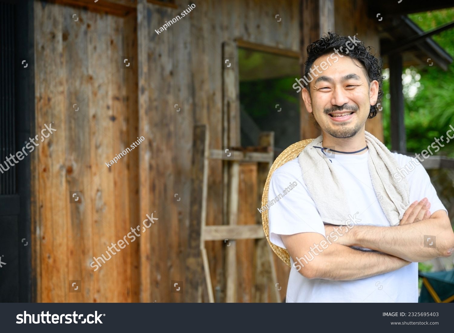 A Japanese farmer stands in the yard of an old house in the Japanese countryside with his arms folded and a smile on his face. The photo shows the upper half of his body, looking at the camera. #2325695403