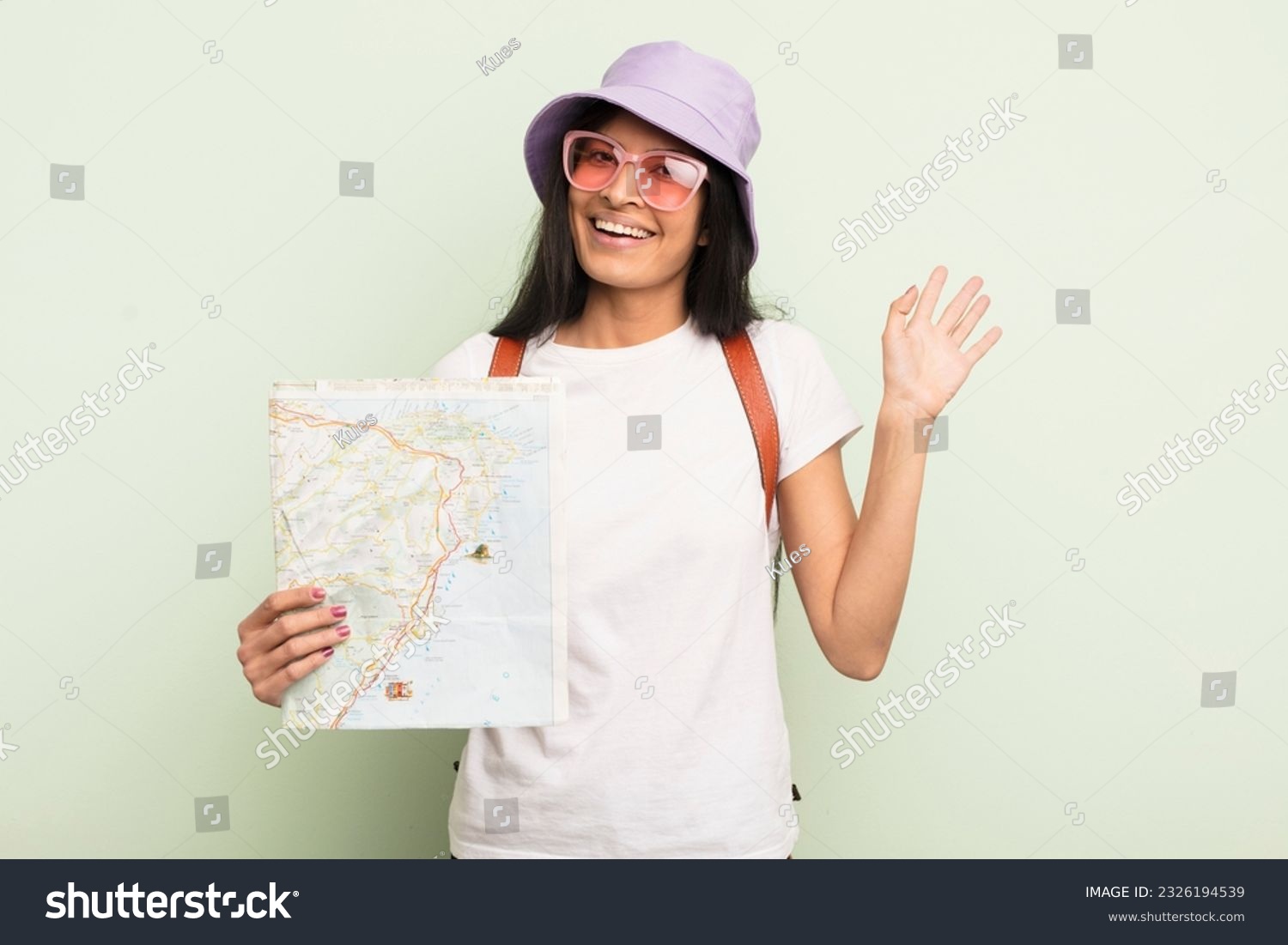 young pretty hispanic woman smiling happily, waving hand, welcoming and greeting you tourist and map concept #2326194539