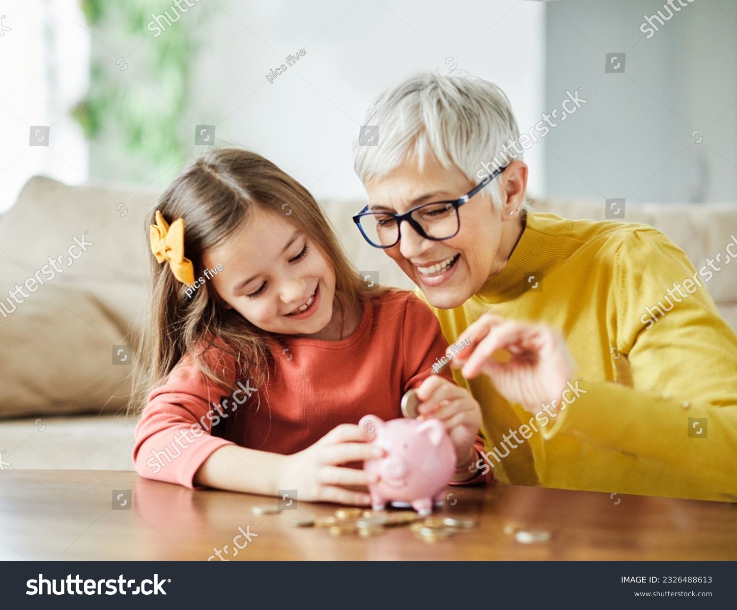 Grandmother and her granddaughter Putting Coin Money In Piggybank  At Home. Personal Savings, Bank Safety And Financial Investments Concept #2326488613