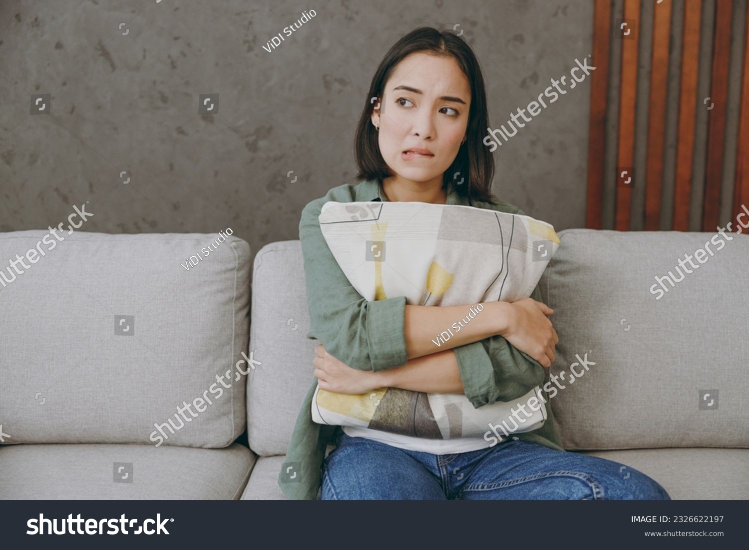 Young sad frustrated upset woman of Asian ethnicity wears casual clothes hugging holding pillow sits on grey sofa couch stay at home hotel flat rest relax spend free spare time in living room indoor #2326622197