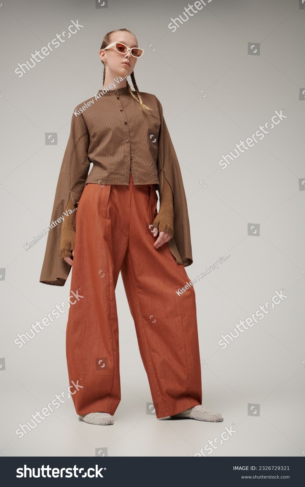 Trends. Haute couture collection. Beautiful fashion model girl posing in a designer brown shirt, knitted gloves and orange wide leg pants. Studio shot on a gray background. Stylish sunglasses. #2326729321