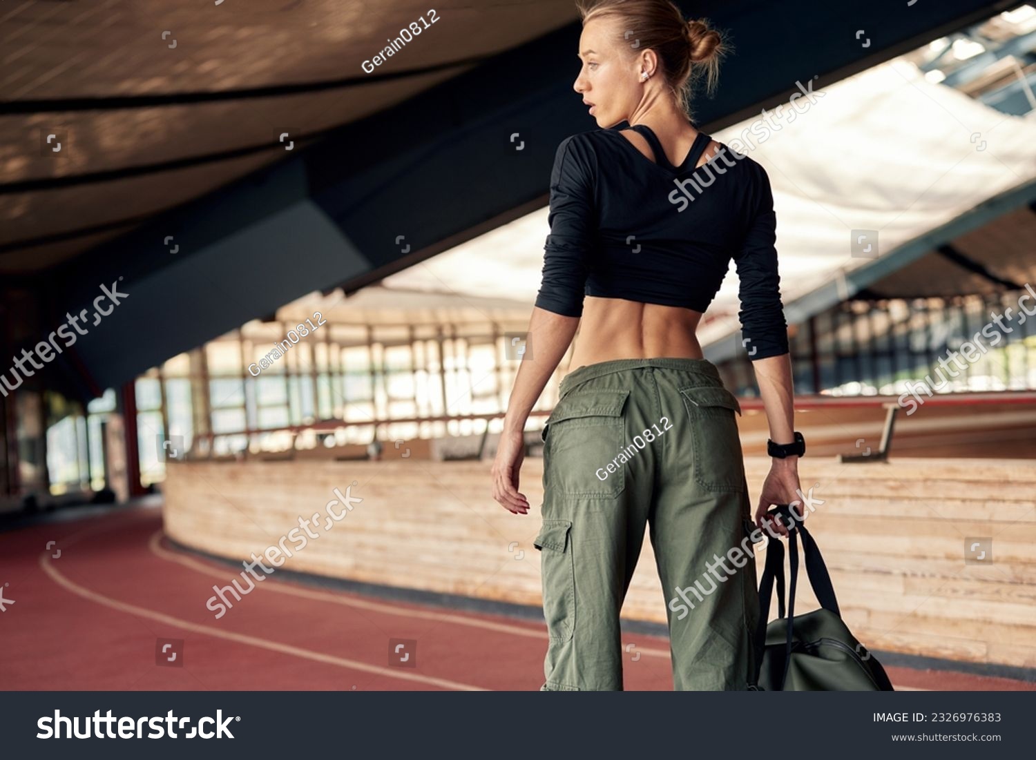 Cute young athlete girl getting ready for a workout at the stadium. Attractive slim blonde in a black top and khaki cargo pants with a sports bag goes to the locker room. Active lifestyle and sports. #2326976383