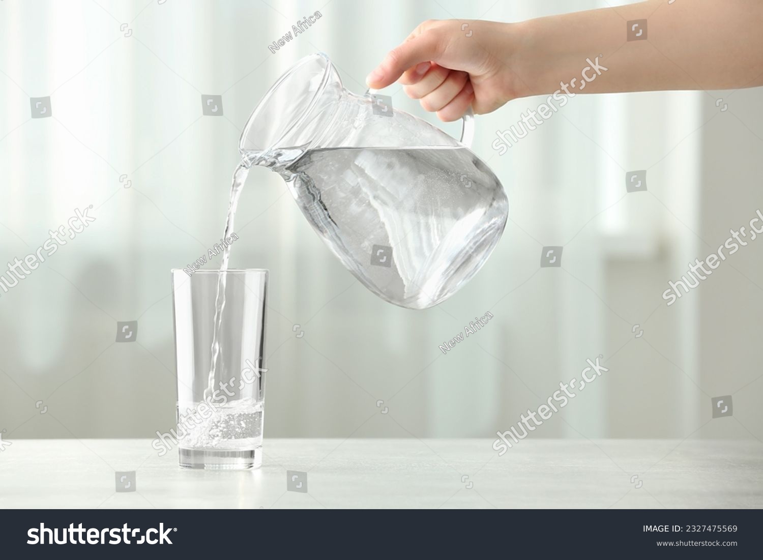 Woman pouring water from jug into glass at white table indoors, closeup #2327475569
