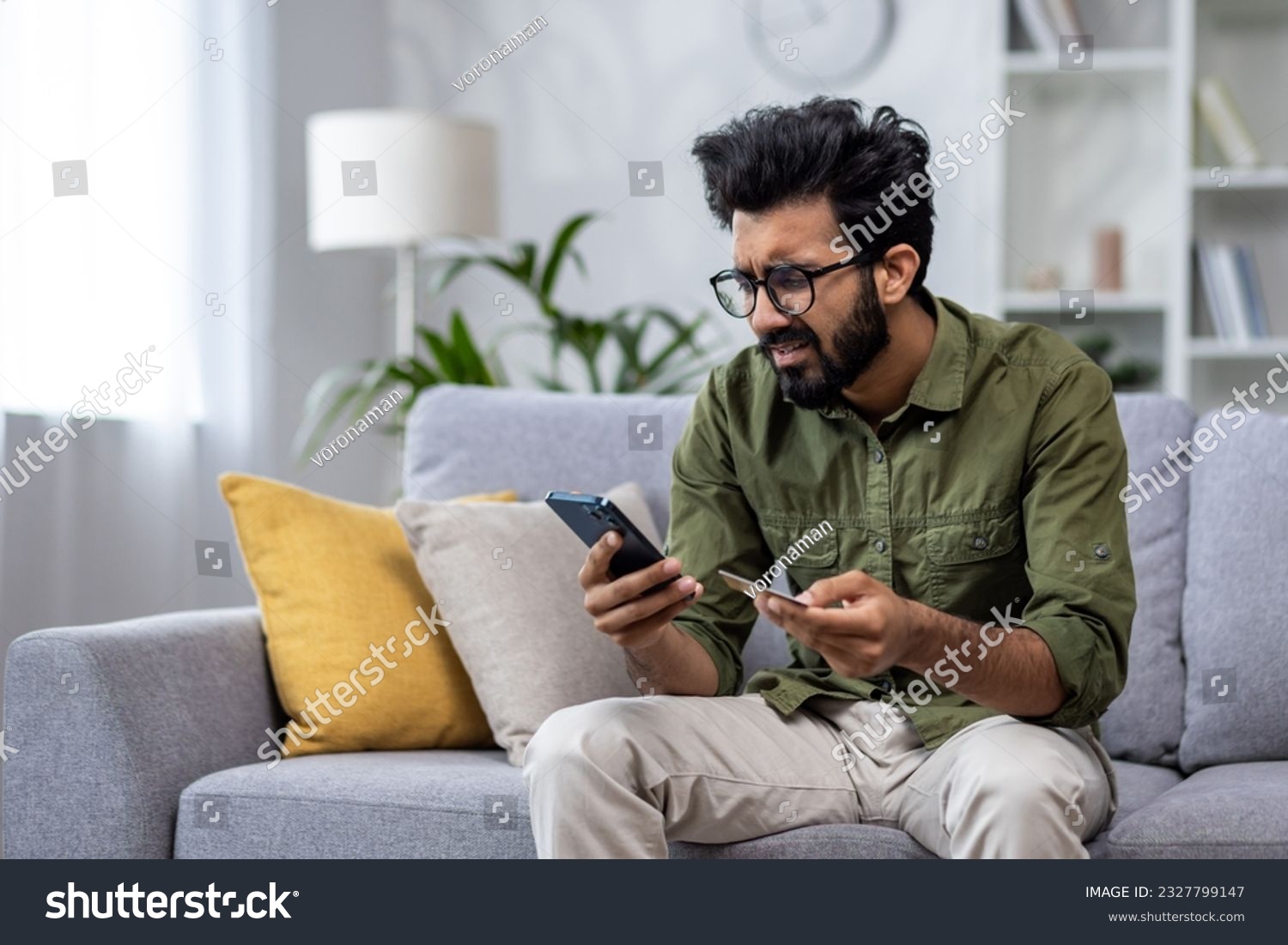 Upset man at home cheated and denied money transfer, hispanic sitting on sofa at home with phone and bank credit card, cheated and disappointed in living room. #2327799147
