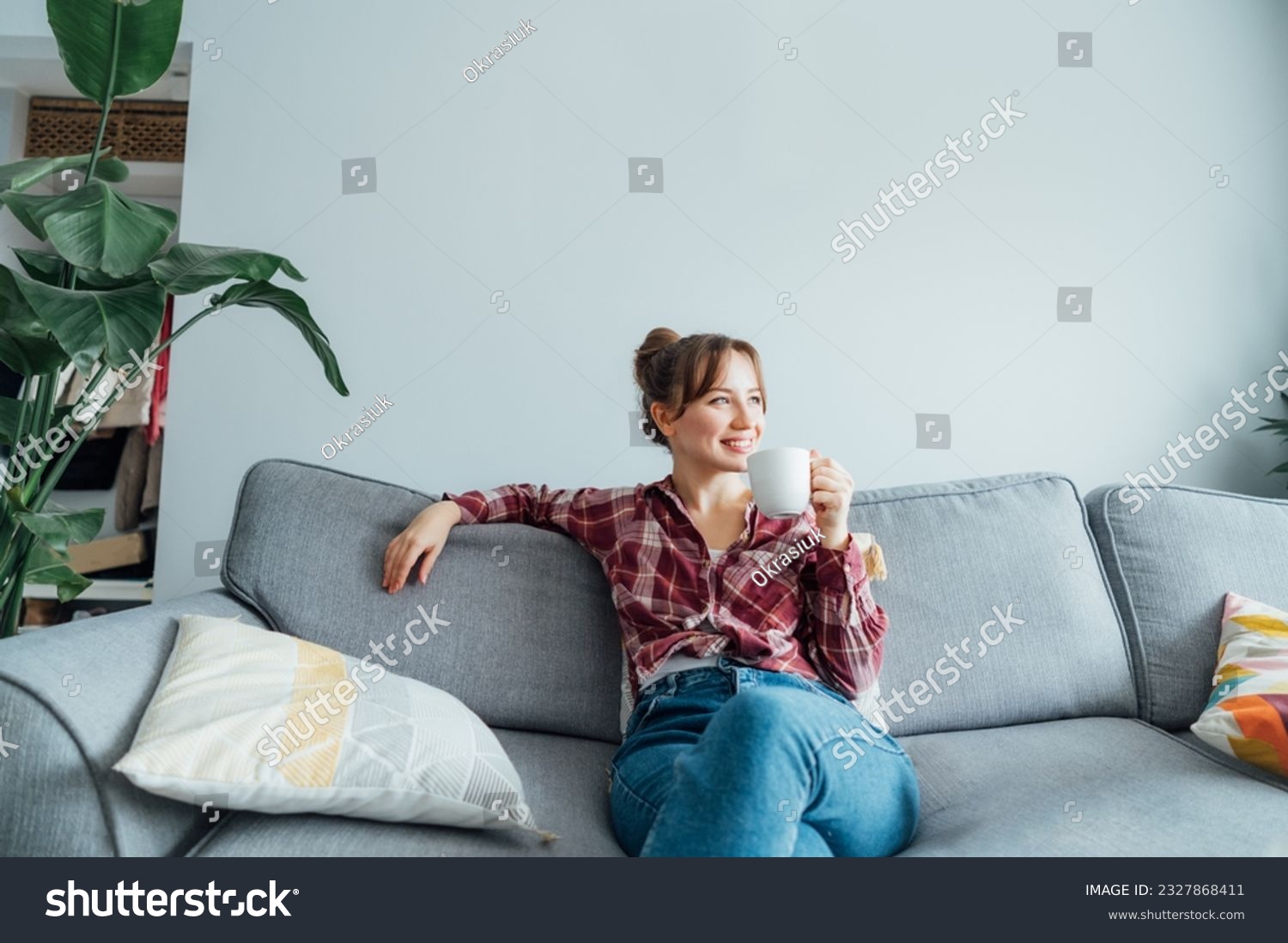 Young smiling woman sitting on sofa and looking away while drinking coffee or tea. Young brunette woman relaxing after housekeeping, home cleaning. Portrait of relaxed female resting at home. #2327868411