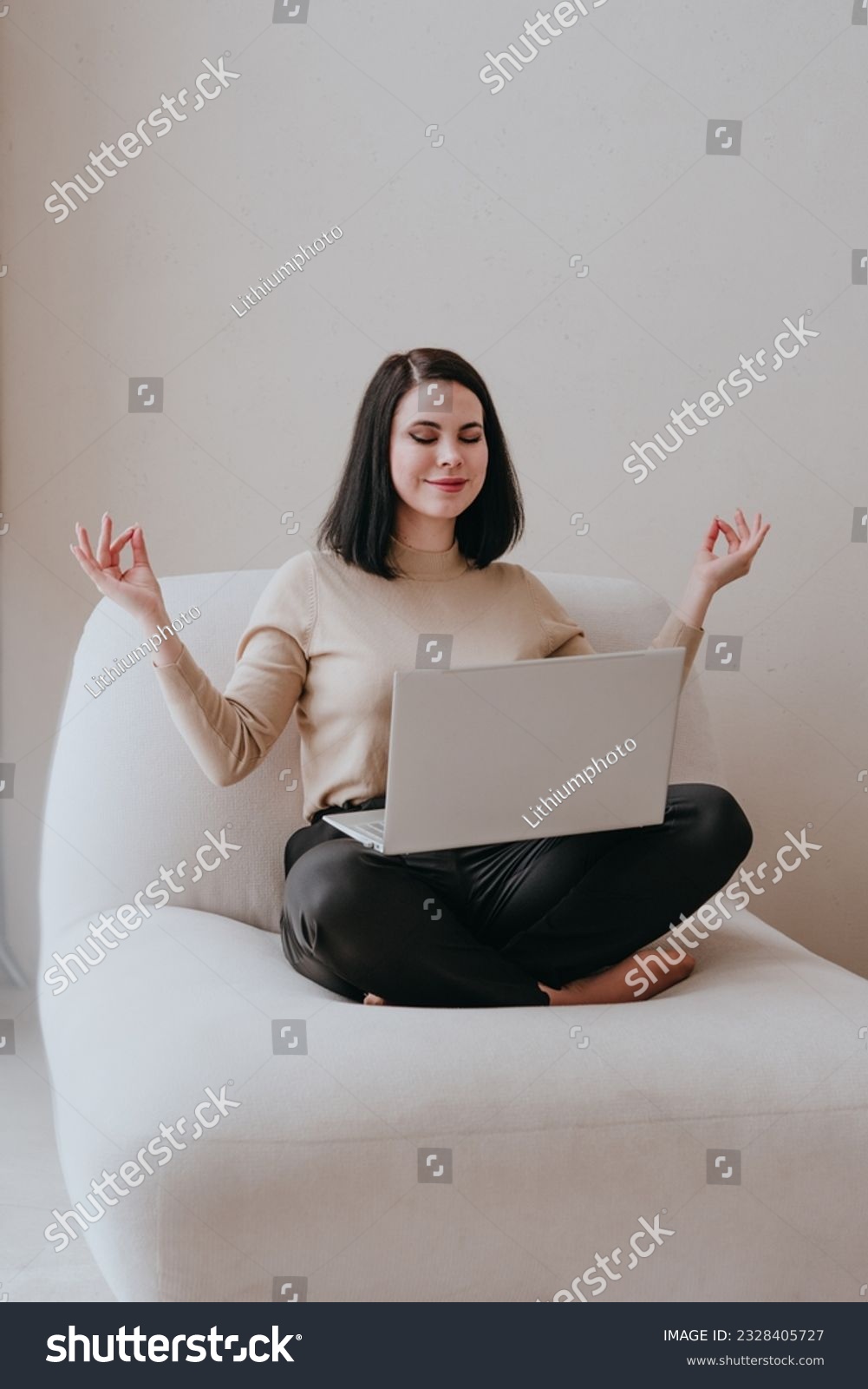 Young smiling brunette woman with closed eyes in business clothes siting barefoot in a lotus position with a laptop indoor. Balance of work and mental health concept. #2328405727