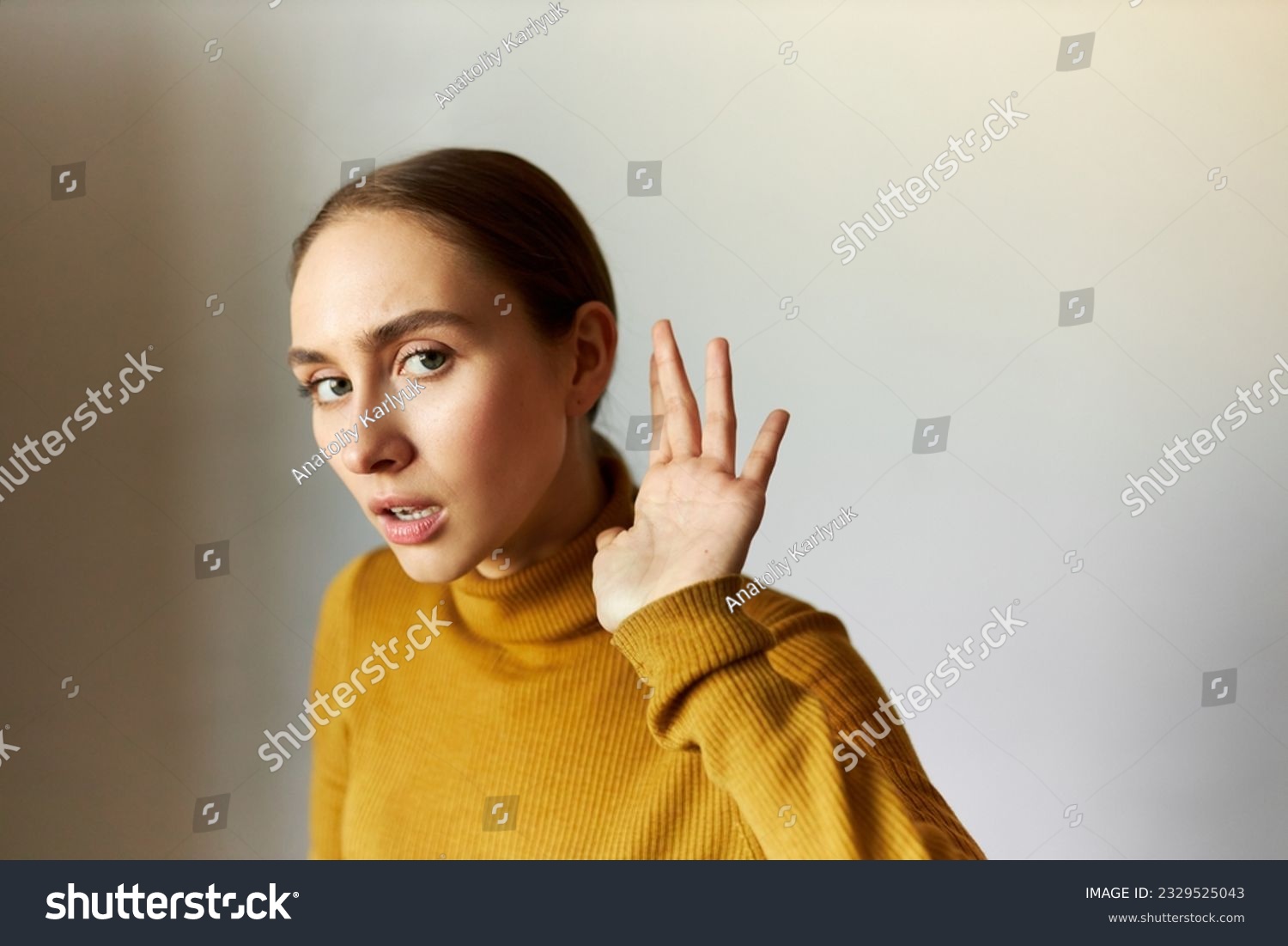 Portrait of concentrated anxious female eavesdropping holding head next to ear, listening carefully to strange sounds, feeling scared, standing against gray studio background with opened mouth #2329525043