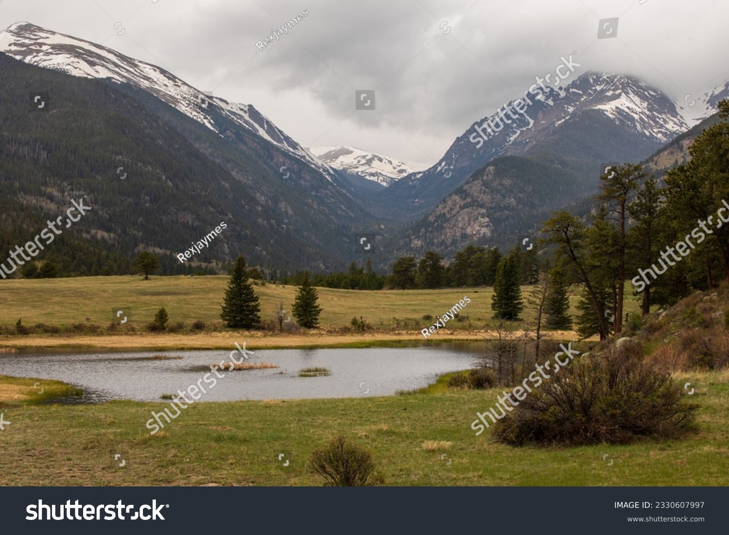 Green nature setting with a pond in foreground and a pale, cloudy sky.  Rocky Mountain pond with reflective water, green grass and dark mountains.  Colorado high country with cloudy sky. #2330607997