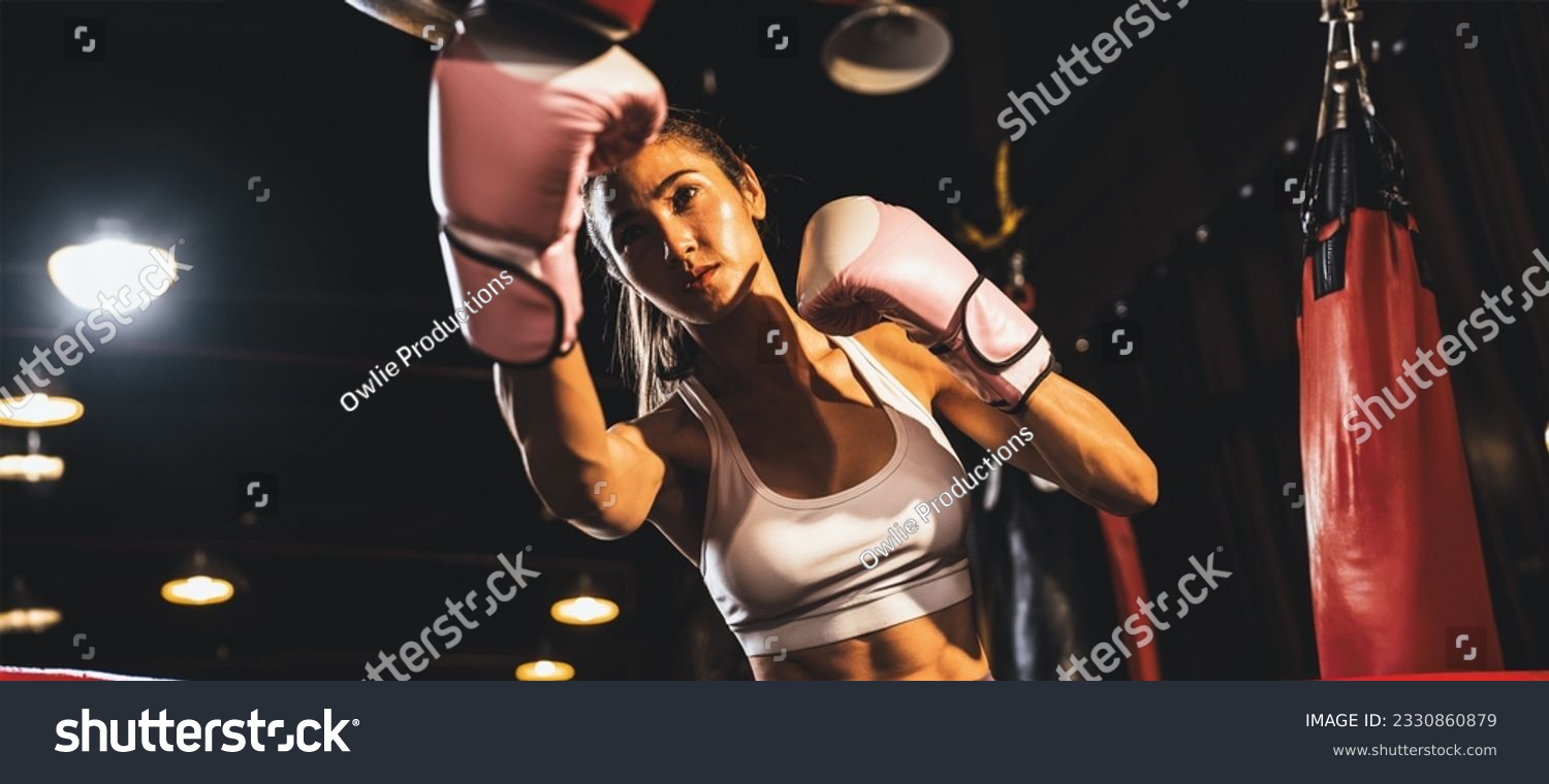 Asian female Muay Thai boxer punching in fierce boxing training session, delivering strike to her sparring trainer wearing punching mitts, showcasing Muay Thai boxing technique and skill. Spur #2330860879