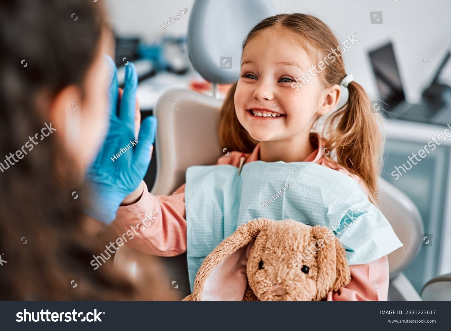 At the doctor's appointment. A candid emotional photo of a child sitting in a dental chair, holding a toy rabbit and cheerfully giving a high-five to the nurse. #2331223617
