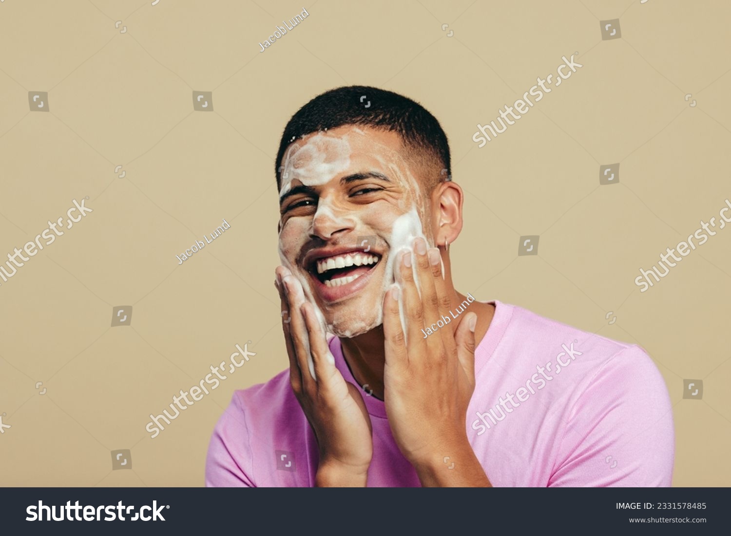 Portrait of a handsome young man indulging in his grooming and self-care routine as he applies face wash in a studio. Youthful man smiling happily while massaging his skin with a facial cleanser. #2331578485