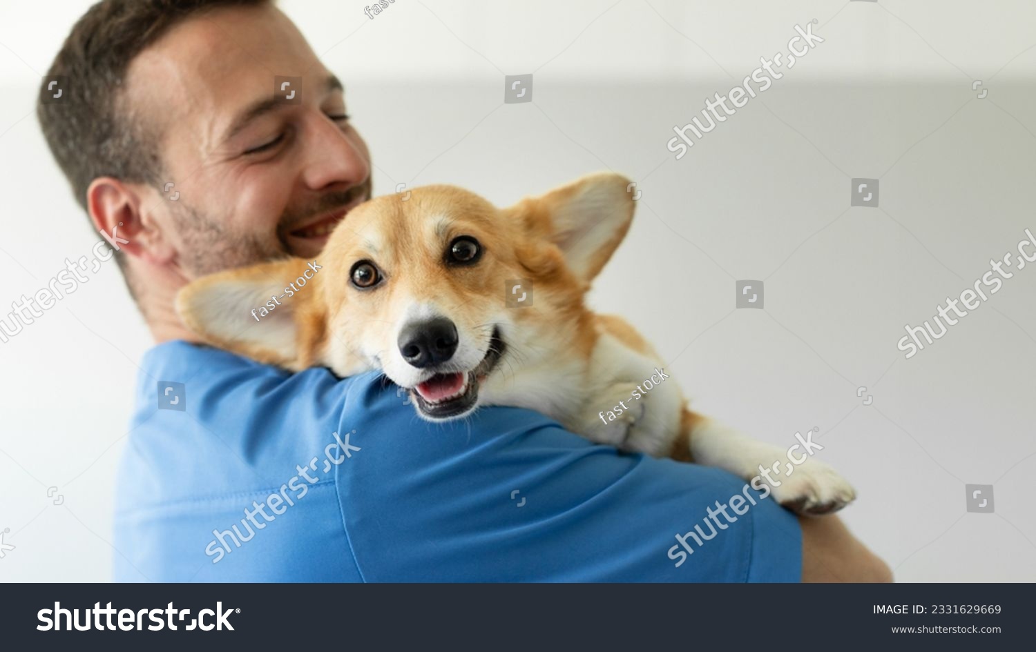 Friendly man vet in blue uniform cuddling, embracing pembroke welsh corgi dog, doctor carrying and playing with little dog after treatment, panorama, empty space #2331629669