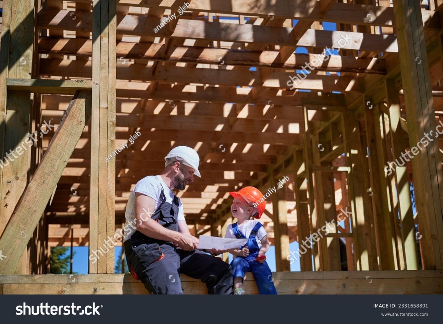 Father with toddler son building wooden frame house. Boys having fun on the edge of balcony, examining the construction plan, wearing helmets and overalls on sunny day. Carpentry and family concept. #2331658801