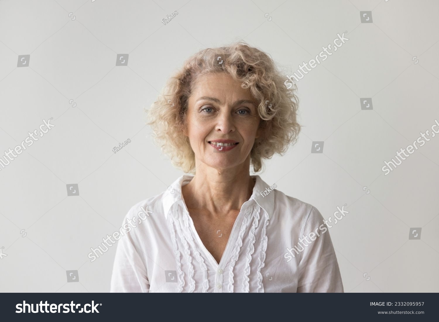 Positive beautiful retired woman standing at white wall background, posing for shooting, looking at camera, smiling. Pretty mature female model with blonde curly hair head shot portrait #2332095957