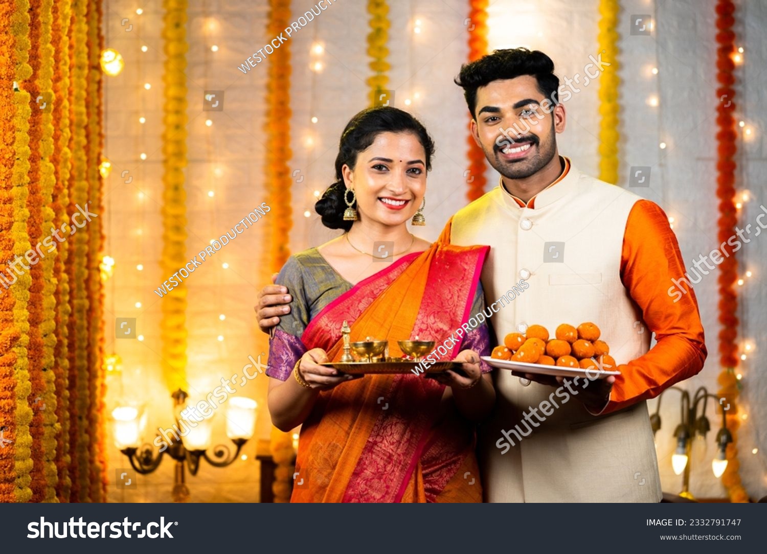 Happy young couple with traditional ethnic wear looking camera by holding sweets and pooja thali or plates - conept of festival celebration, rituals and cultural beliefs #2332791747