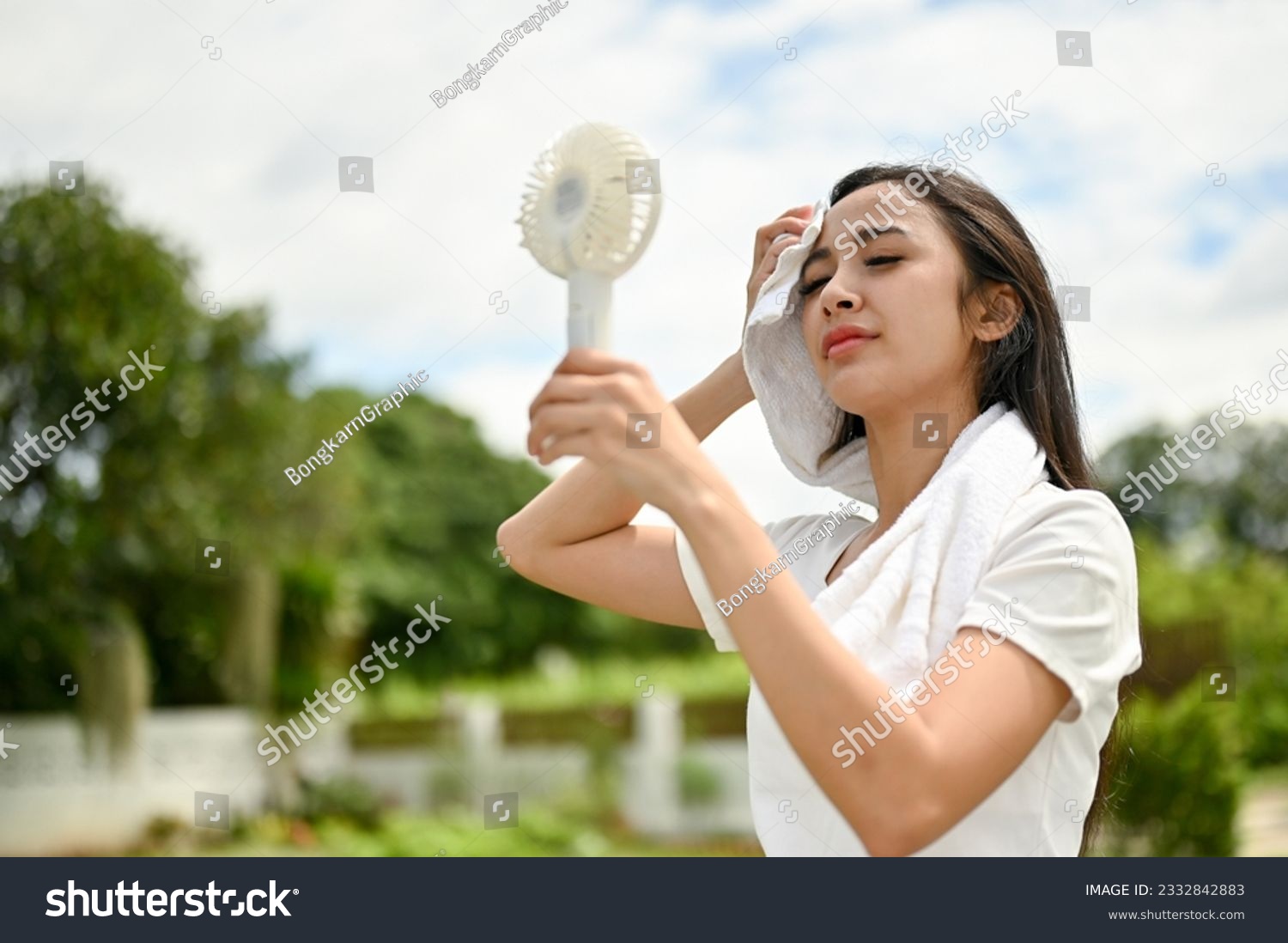 An exhausted and sweaty Asian woman in sportswear using a portable handy fan, feeling hot and tired after a long run at the park on a sunny day. #2332842883