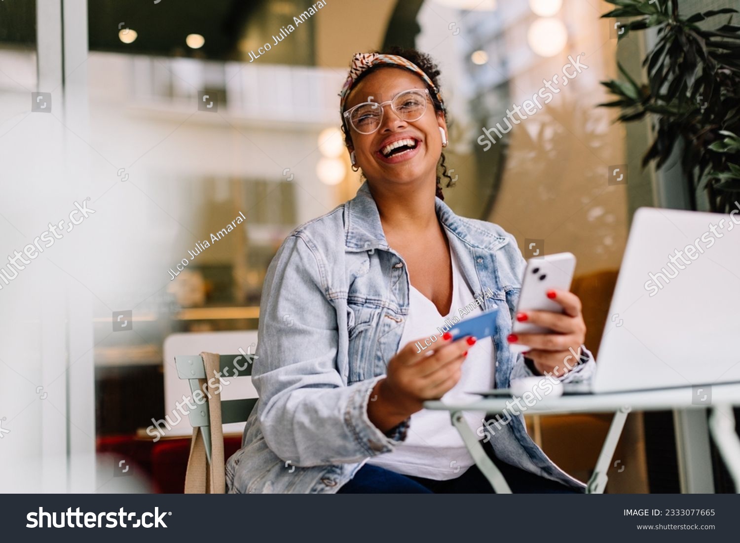 Happy woman sitting in a café and using her smartphone for online shopping, paying with a credit card. Young woman enjoying the convenience of mobile banking and digital transactions. #2333077665