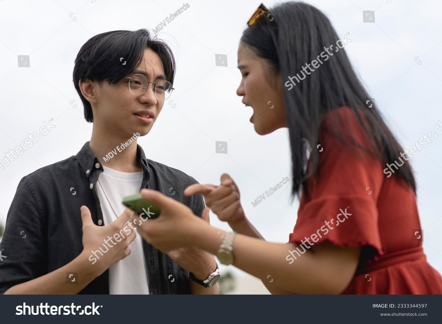 A young asian woman confronts her cheating boyfriend over a extramarital relationship with another girl on his phone. A man taken aback and surprised after getting caught red-handed. #2333344597
