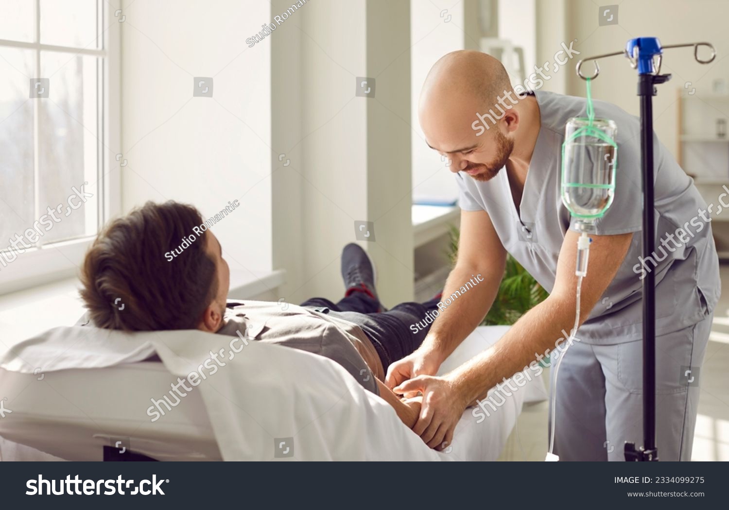 Nursing staff at clinic or hospital gives intravenous vitamin drip or medicine infusion to patient. Nurse personnel inserts venous IV line needle in arm vein of adult man lying on bed in medical ward #2334099275