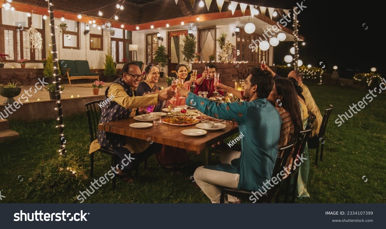 Big Indian Family Celebrating Diwali: Family Gathered Together on a Dinner Table in a Backyard Garden Full of Lights. Group of Adults Having a Toast and Raising Glasses on a Hindu Holiday #2334107399