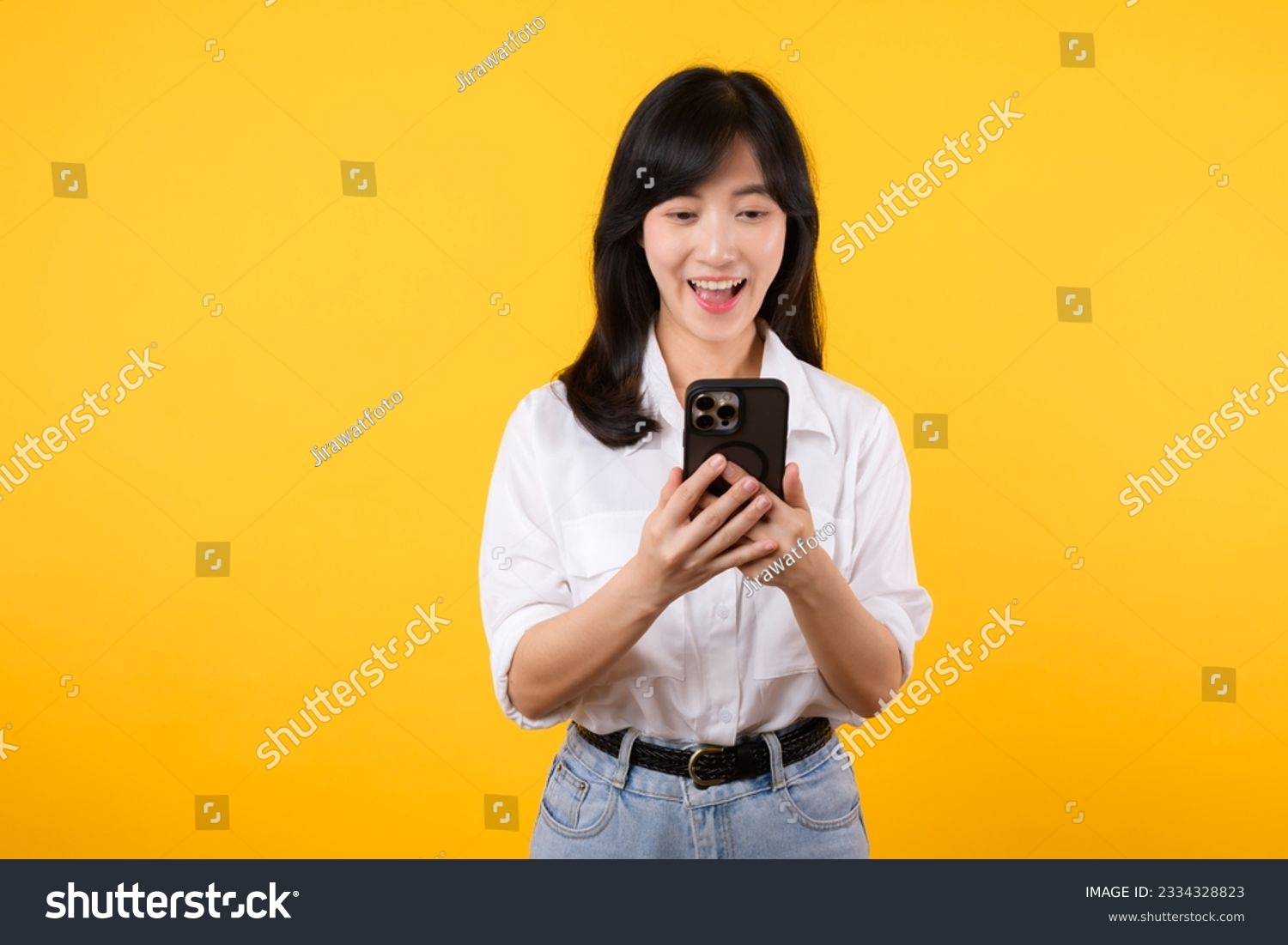 Surprised asian young woman using mobile phone with positive expression, smiles broadly, dressed in white shirt and standing isolated on yellow background. Happy adorable glad woman rejoices success. #2334328823