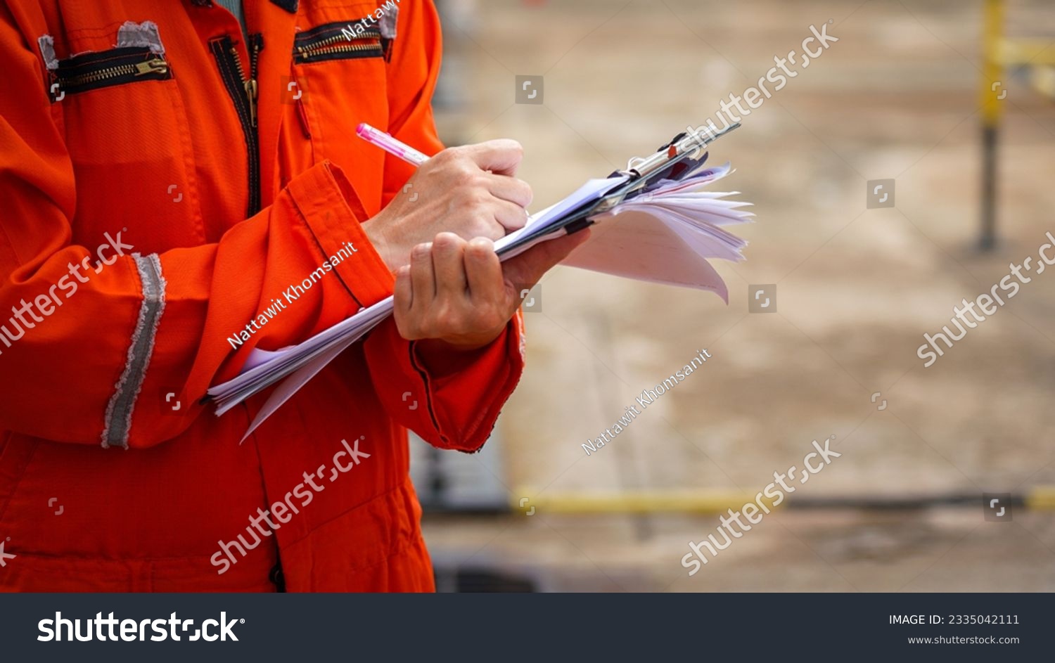 Action of a safety officer in full PPE coverall is writing note on paper document during perform safety audit at construction worksite. Industrial expert working scene. Selective focus. #2335042111