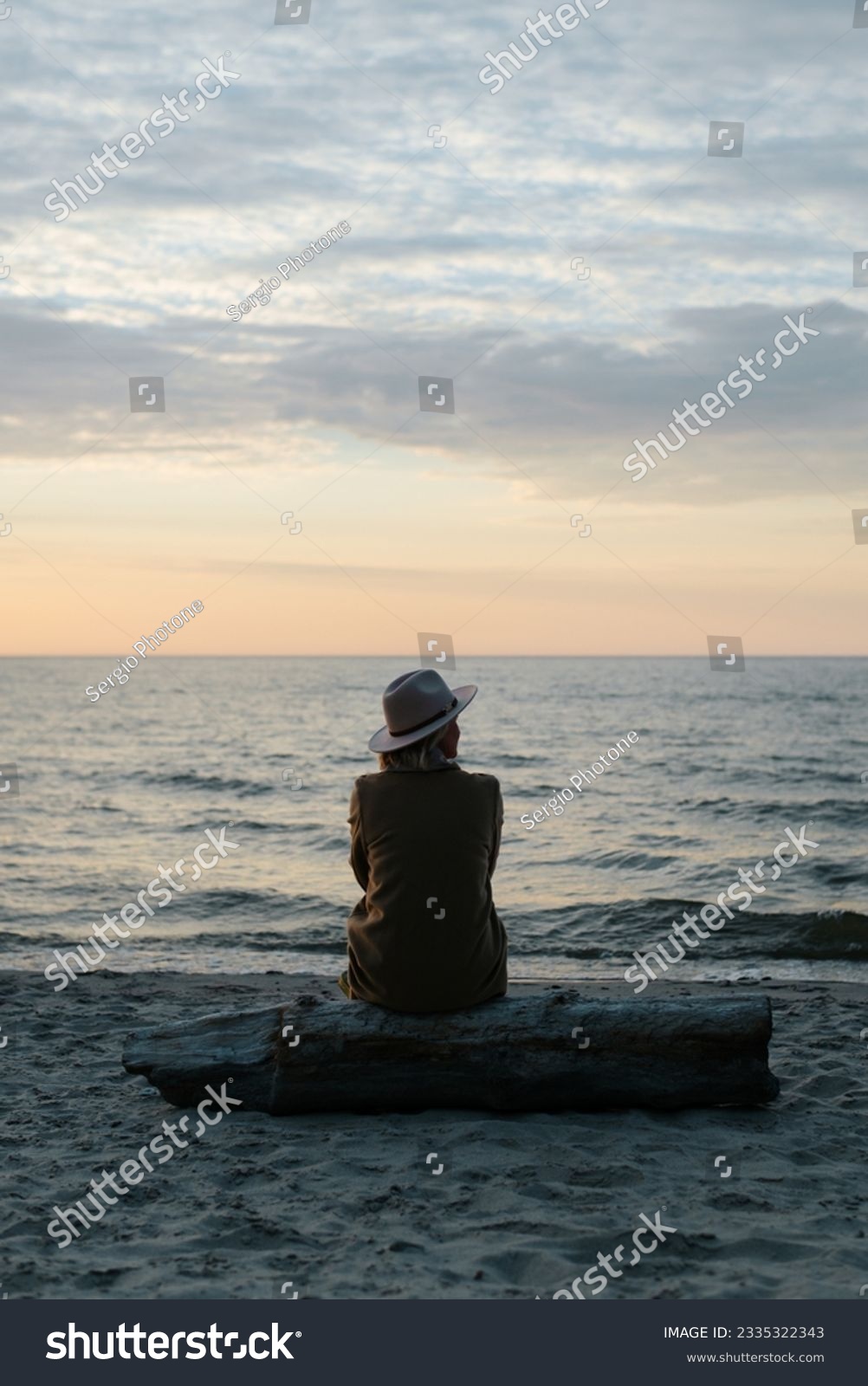 Back view of pensive lonely woman in coat and hat sitting on log on seashore looking away at sea in evening. #2335322343