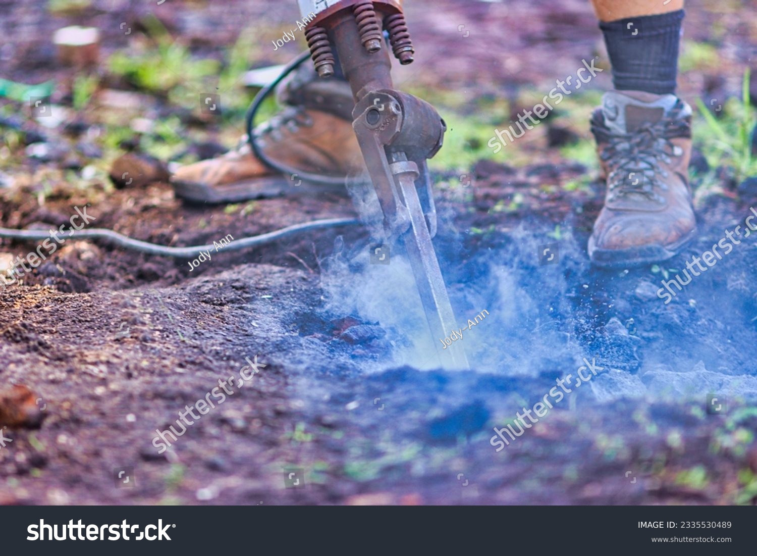 man wearing work boots using a jackhammer that is breaking up rocks and sending dust and debris into the air.   #2335530489