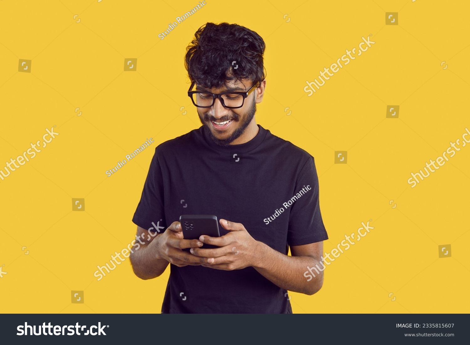 Young joyful Indian man chatting with phone and reading SMS with smile using instant messengers and social networks standing in yellow studio. Mobile technologies, internet communications #2335815607