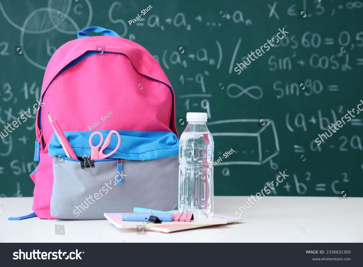 Colorful school backpack with bottle of water and stationery on white table near green chalkboard #2336631305