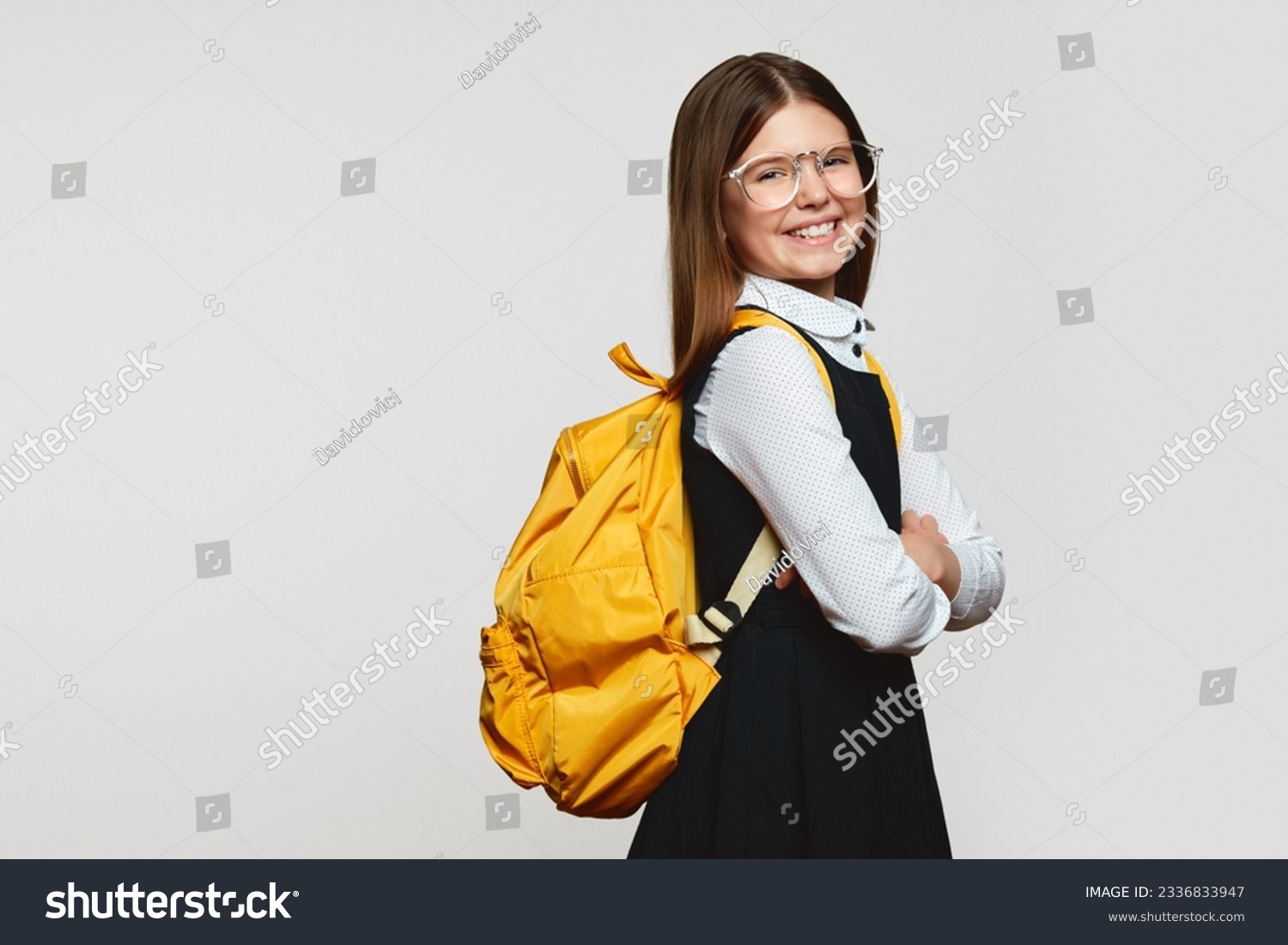Side view of happy schoolgirl wearing glasses and yellow backpack holding crossed hands while smiling against white backdrop with free space for text #2336833947