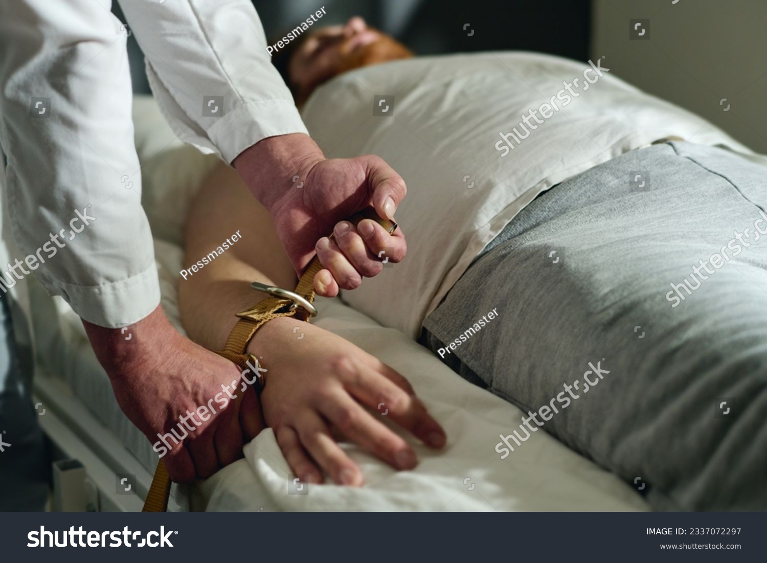 Hands of male doctor of mental hospital binding patient with belts to bed while bending over him during medical treatment course #2337072297