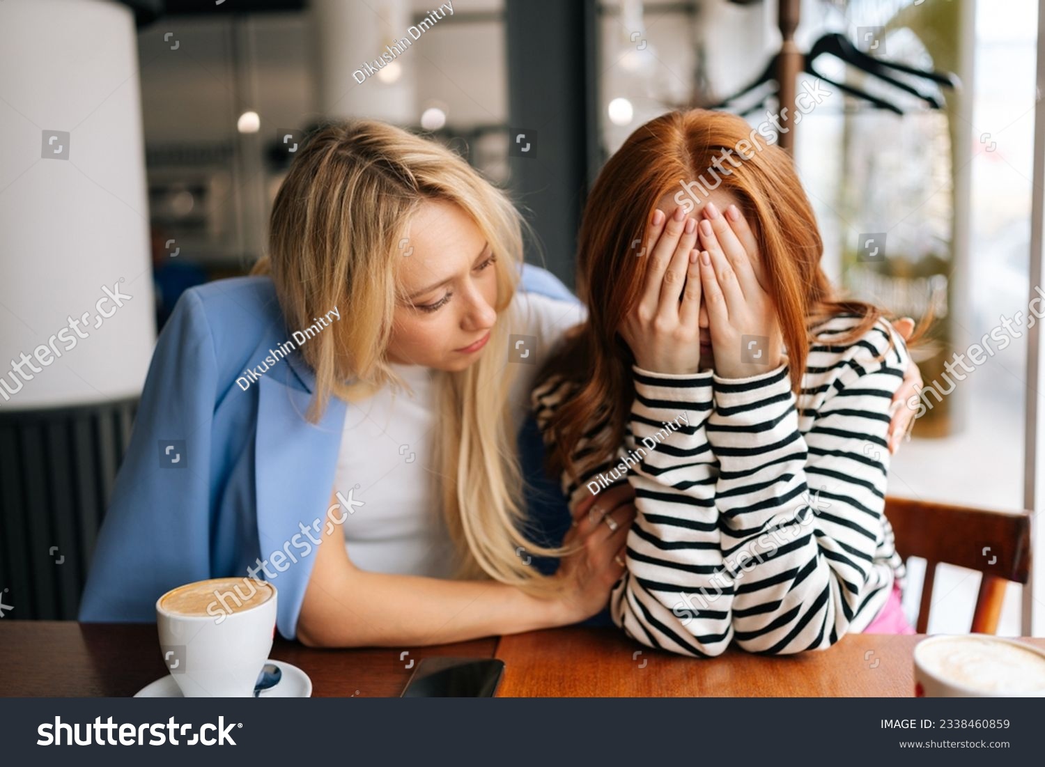 Portrait of displeased young woman and best female friend trying to comfort and cheer up sitting together in cafe by window. Unhappy redhead lady covering face with hands and crying. #2338460859