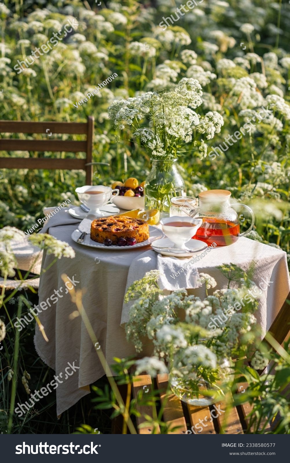 Cozy tea party in the garden with white porcelain cups, teapot, candle, flowers, homemade sweet cherry pie. Wooden furniture, elegant table decor. Romantic atmosphere, candles, linen tablecloth #2338580577
