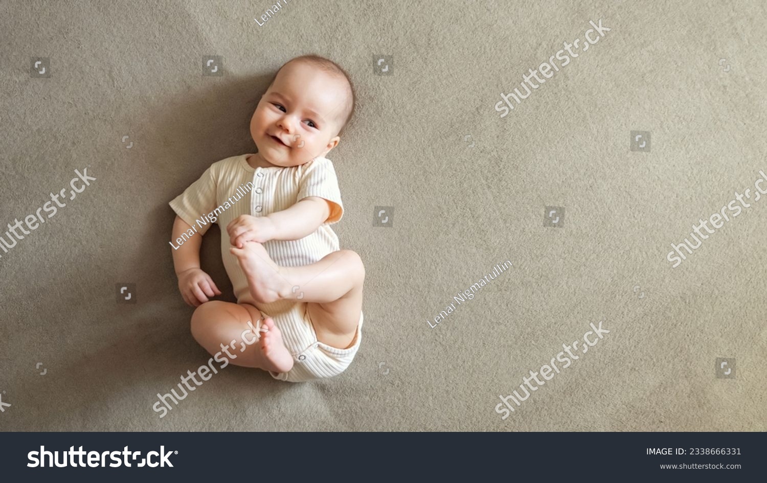 Infant lying on brown bed smiling with joy. Baby girl with delighted expression plays touching toes with hands and relaxing on bed, copyspace #2338666331