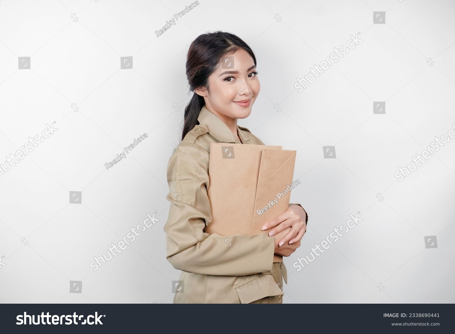 A smiling young beautiful Asian civil worker with brown uniform bringing document. Indonesian PNS. #2338690441