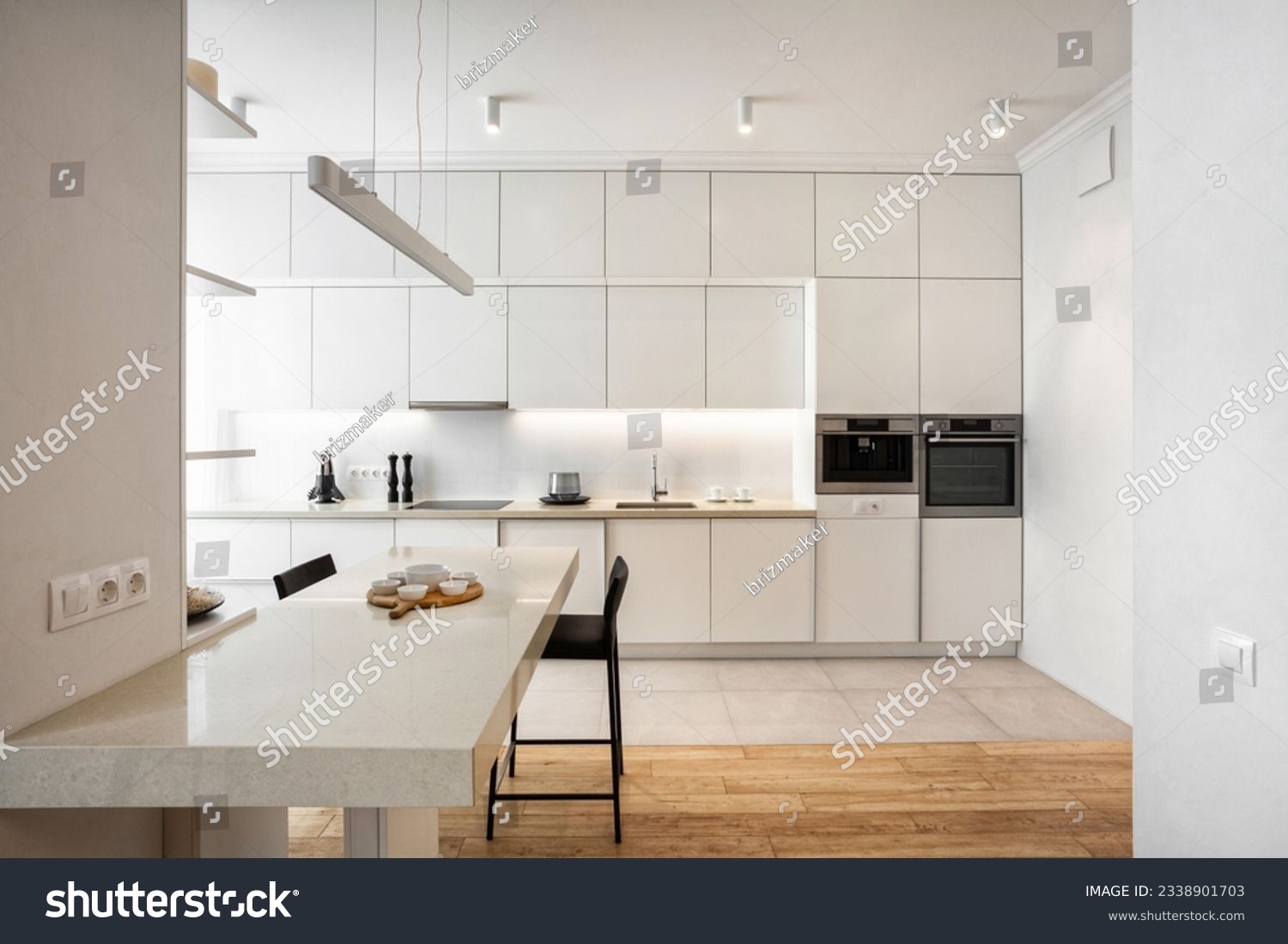 Front view of light minimalistic kitchen with white facade, built-in appliances and marble table. Stylish kitchenware on countertop. Well organized space in new apartment #2338901703